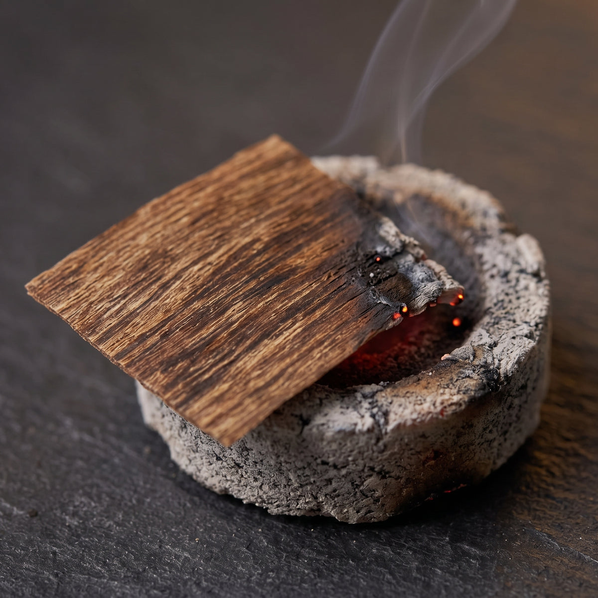 A single Vietnamese oud incense chip smoldering and releasing smoke on a round lit charcoal piece.